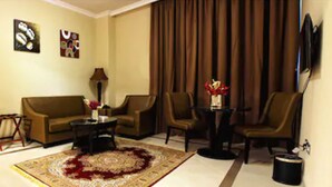 A living room with a sofa, two armchairs, a coffee table, and a TV. There is a large window with curtains, a small table with a lamp, and a decorative rug. - Paragon Hotel Apartments (Abu Dhabi)