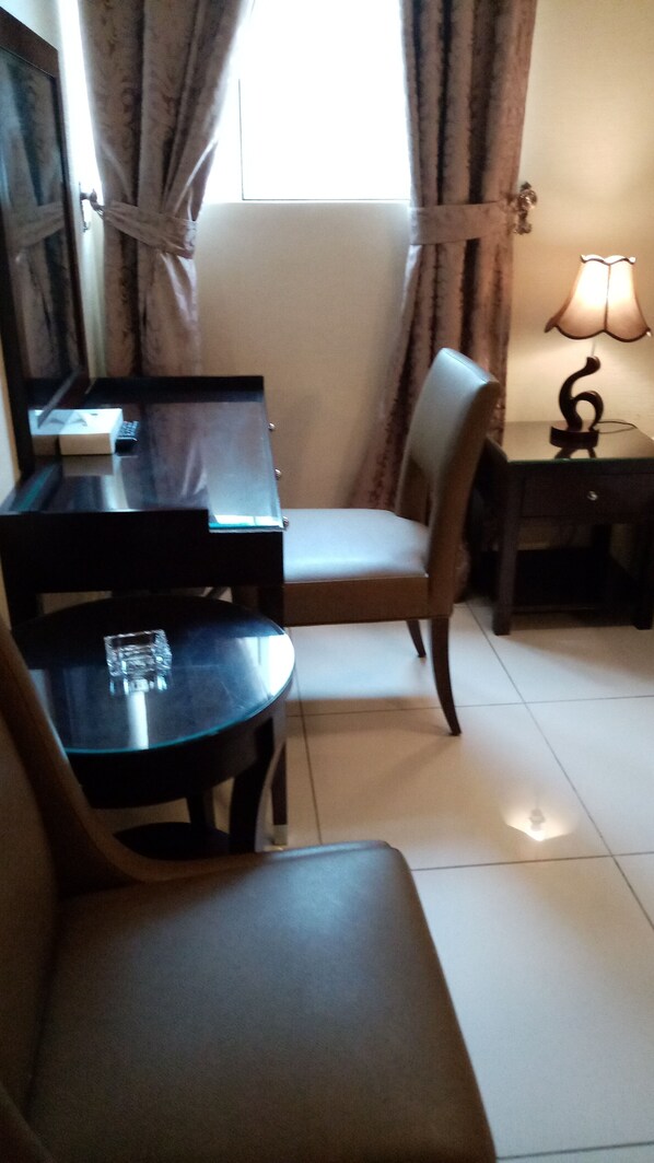 A room with a dining table, a chair, a small table with a lamp, and a window with curtains.