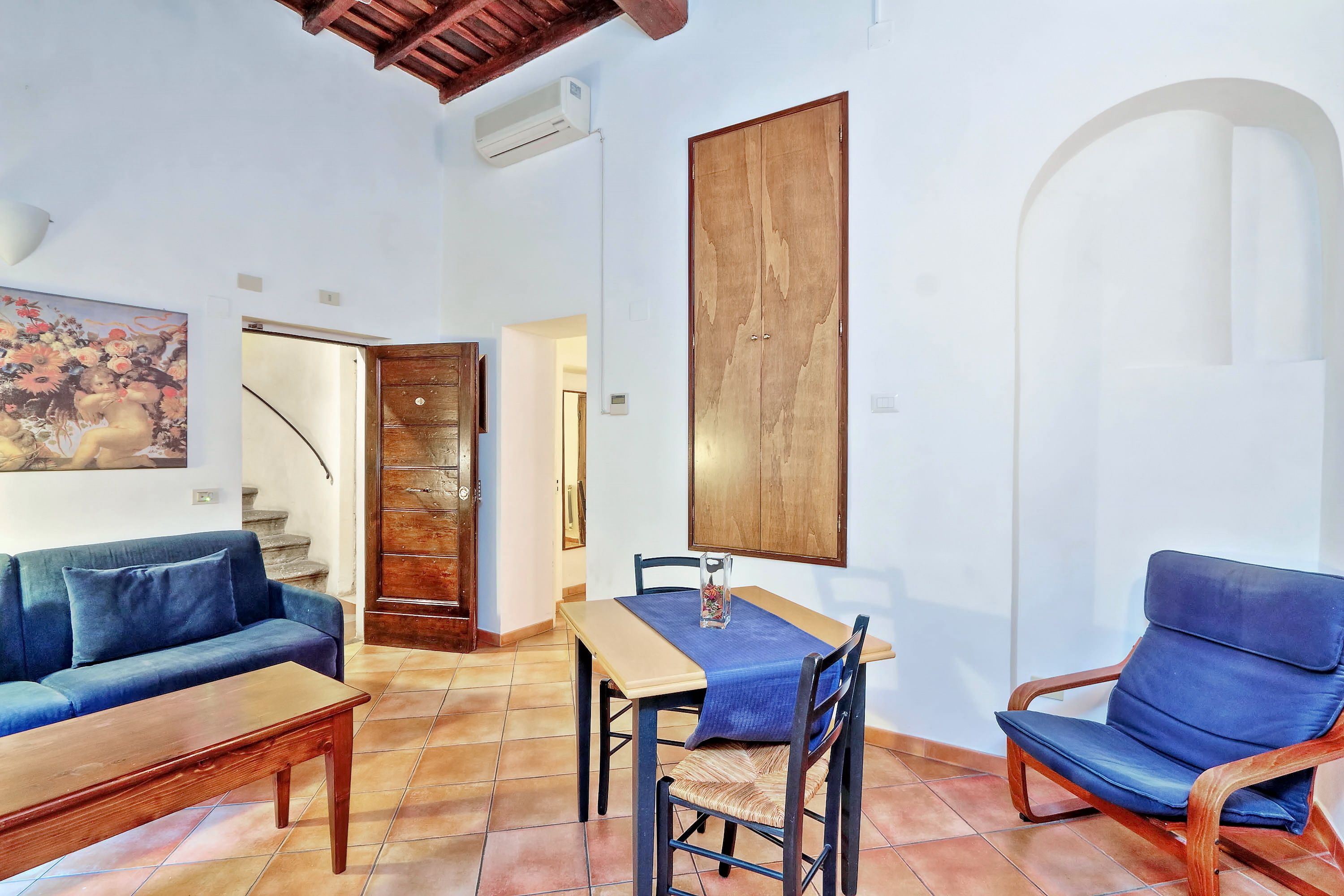 A living room with a sofa, a coffee table, a dining table with chairs, a painting on the wall, and a wooden wardrobe.