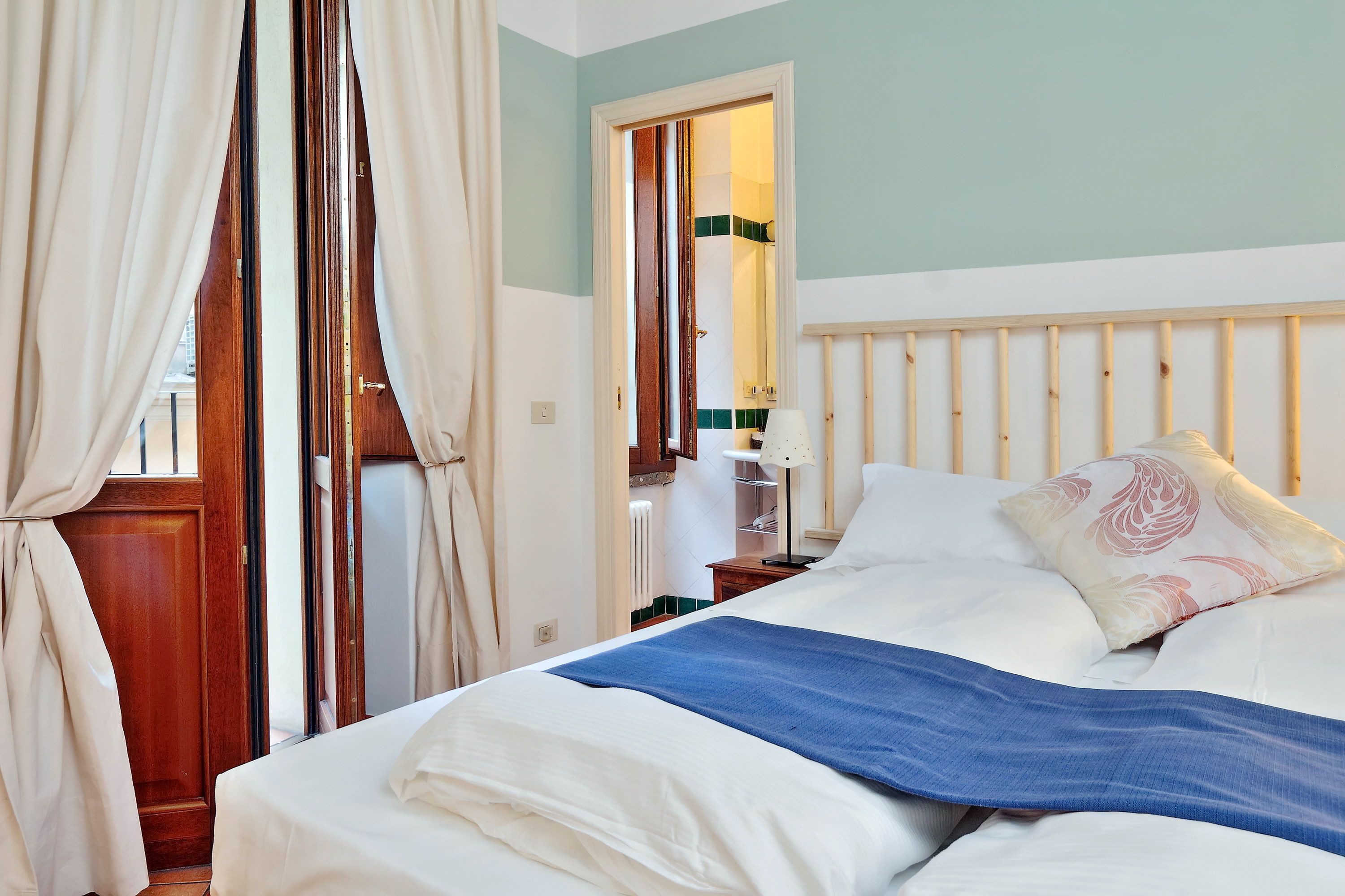 A neatly made bed with white and blue bedding, a wooden headboard, and a window with curtains.