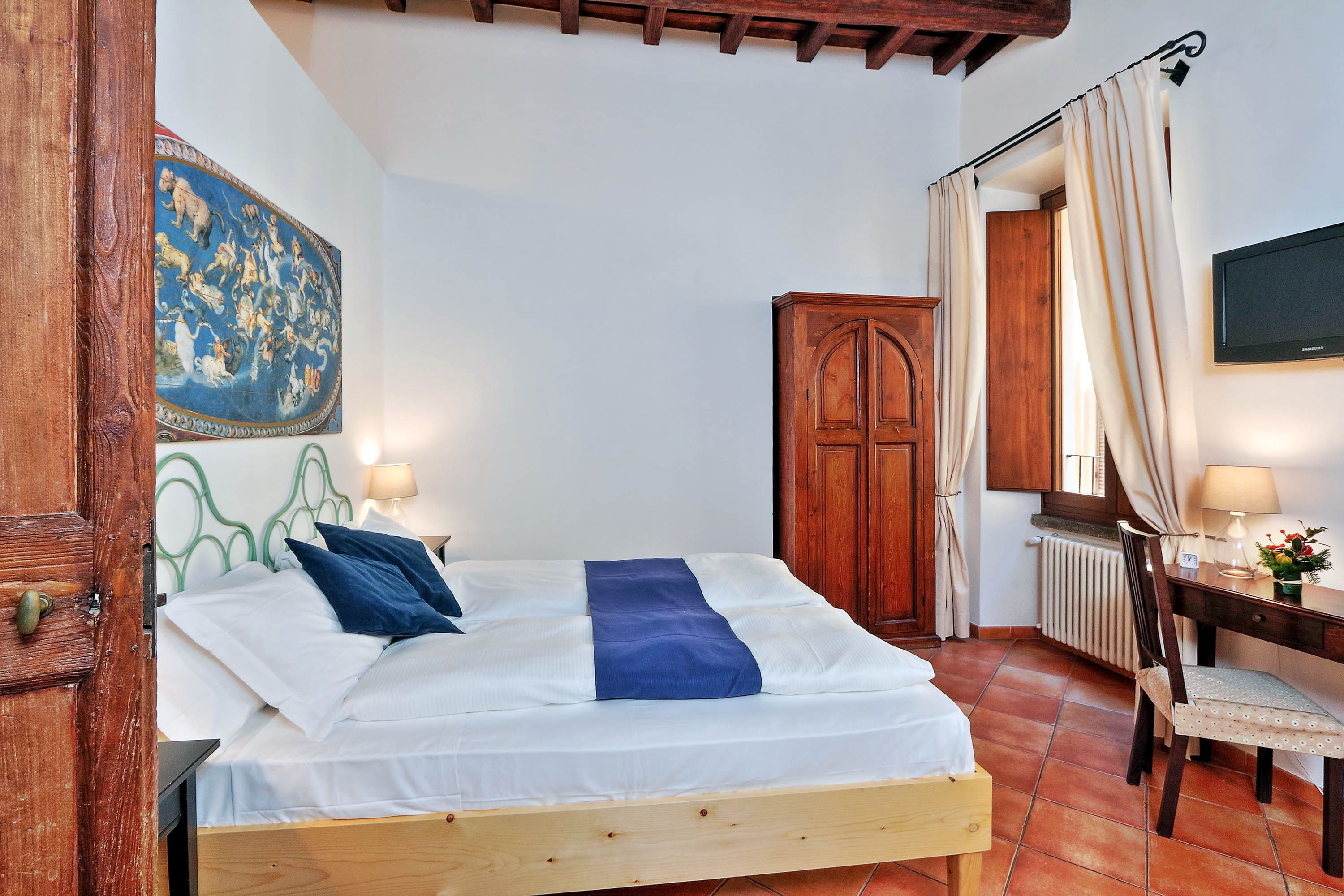 A bedroom with a wooden bed, a desk with a chair, a radiator, and a television on the wall.
