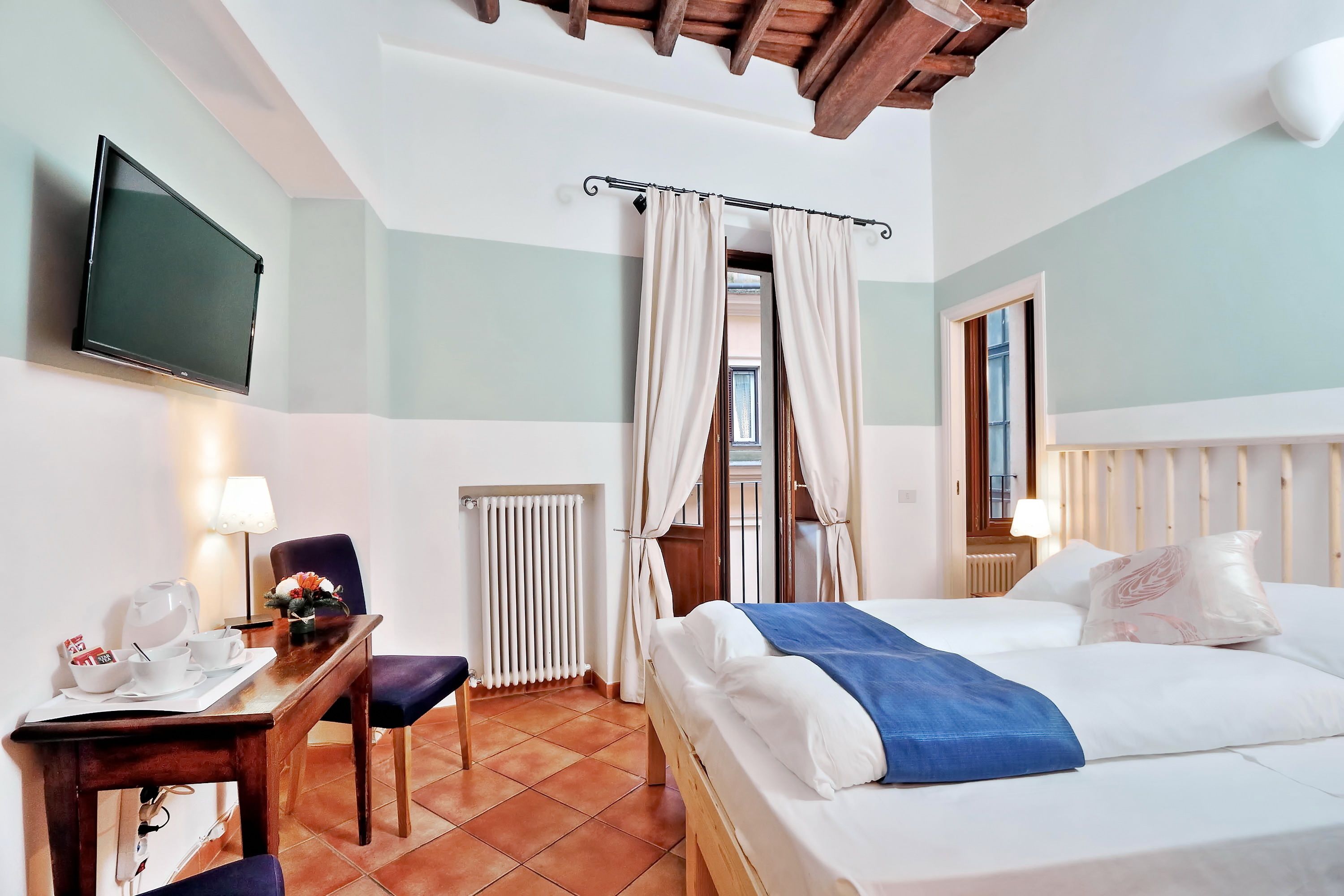 Habitación de hotel con cama, escritorio y silla, televisor montado en la pared y ventana con cortinas.