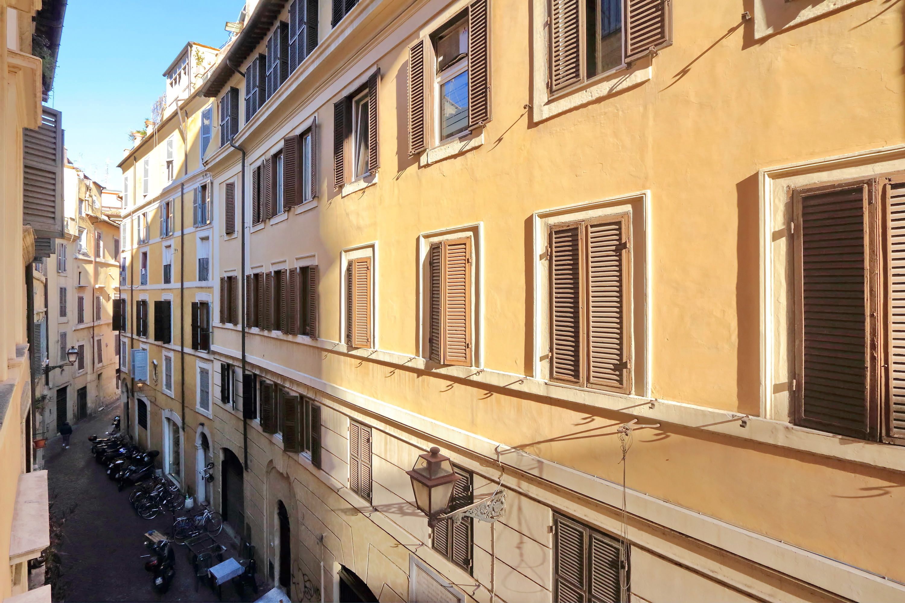 Una stretta via fiancheggiata da edifici, con balconi dotati di persiane in legno e una fila di scooter parcheggiati.