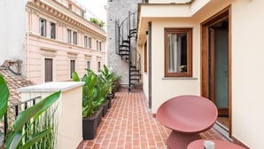Un corridoio esterno stretto con pavimento in piastrelle rosse, piante in vaso, una sedia rossa e una scala che conduce a un livello superiore.