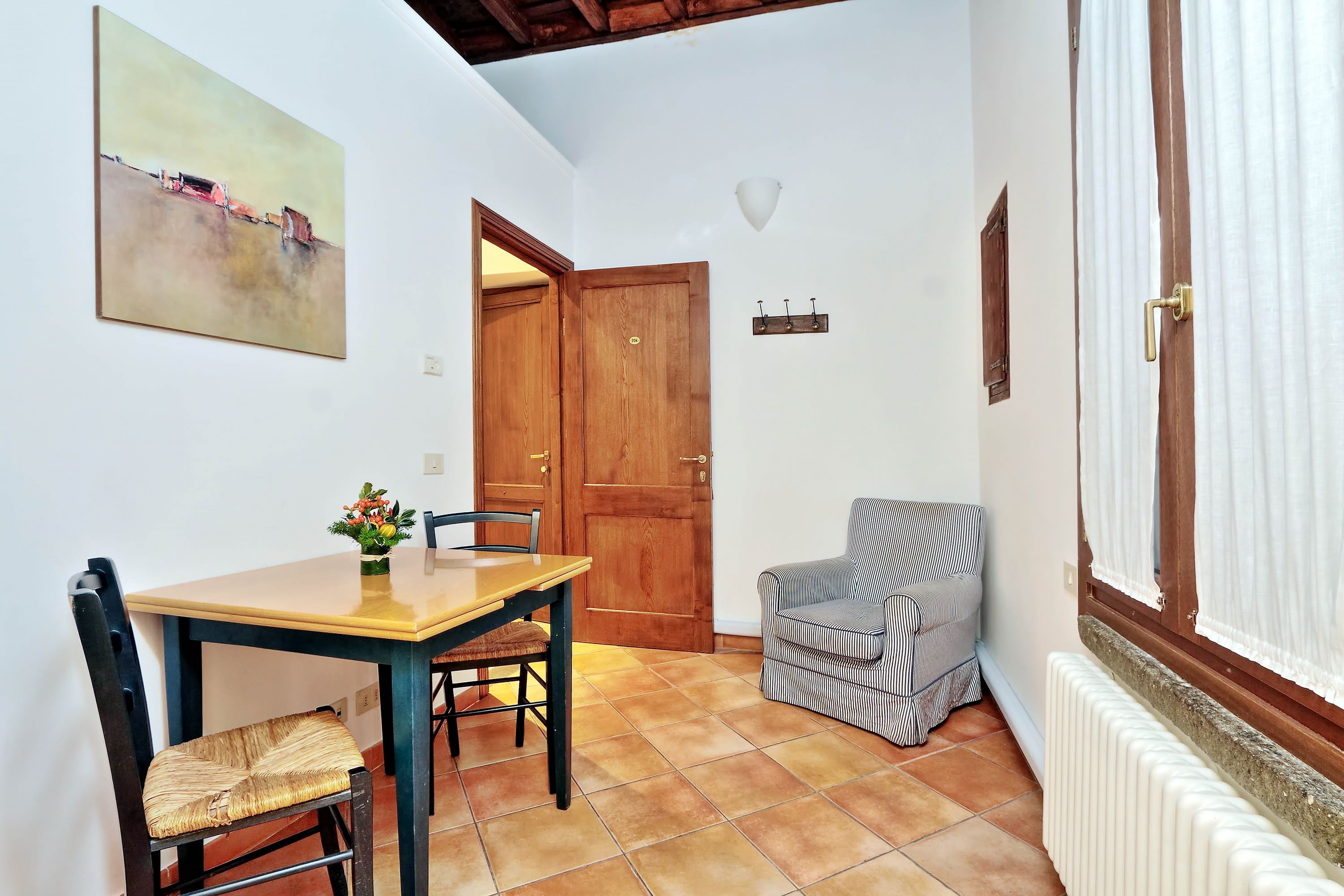 A dining area with a wooden table and chairs, a painting on the wall, a wooden door, a radiator, and a window with curtains.