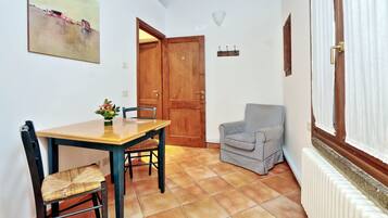 A dining area with a wooden table and chairs, a painting on the wall, a wooden door, a radiator, and a window with curtains.