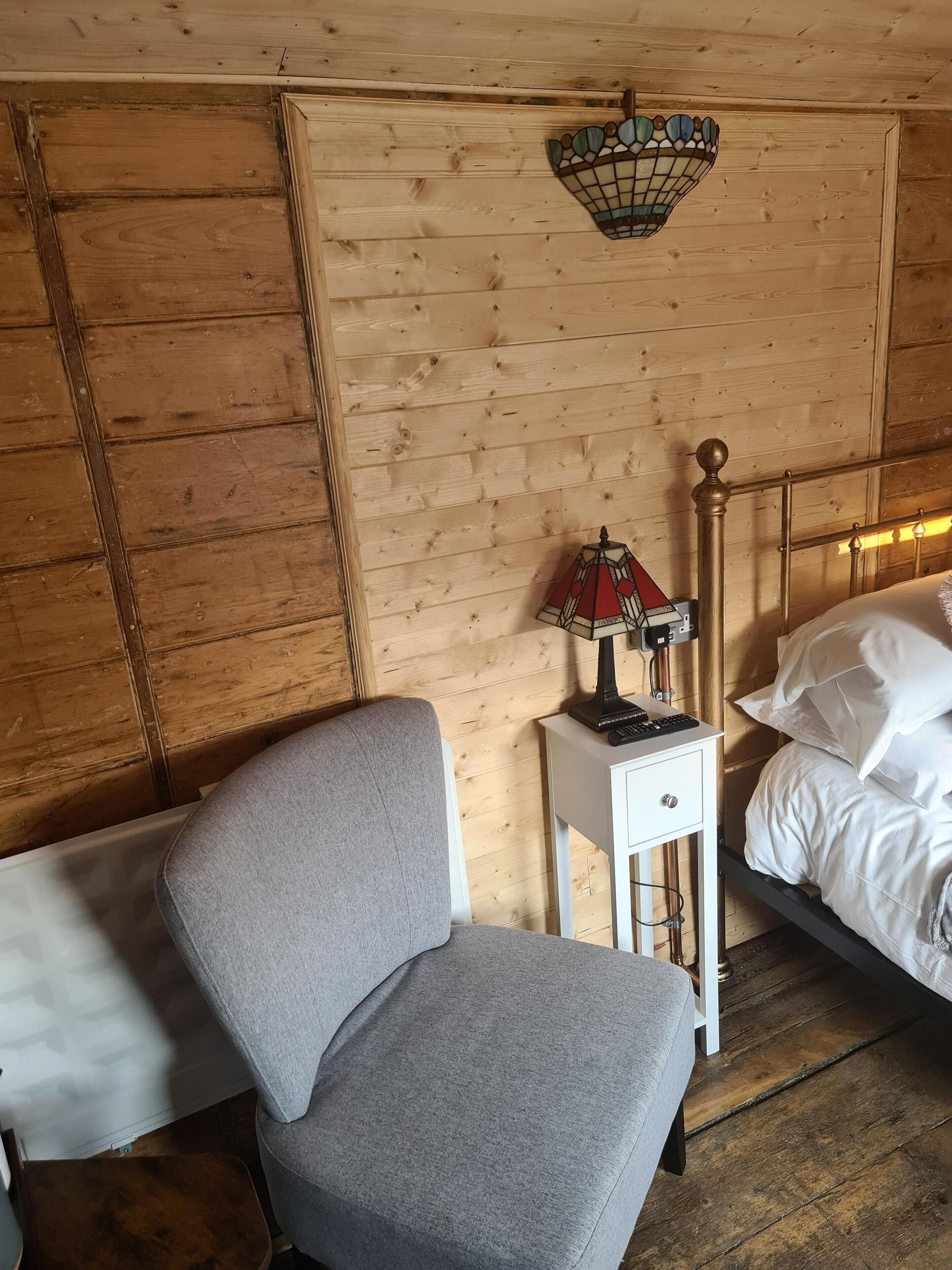 A wooden-paneled room with a bed, a nightstand with a lamp, and a grey chair.
