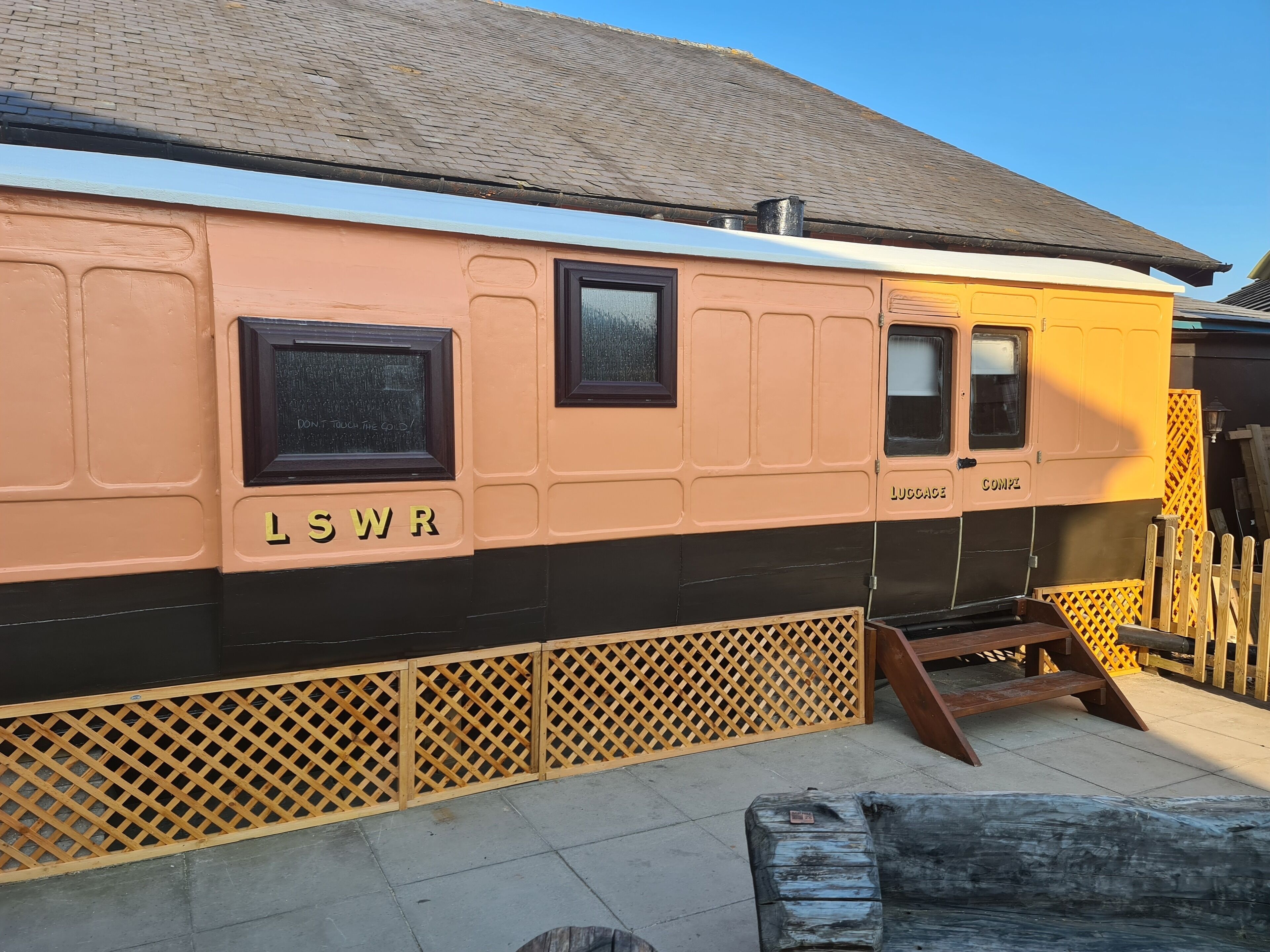 Un vagón de tren antiguo con letreros que dicen "L S W R" y "LUCCAGE", una cerca de madera con celosía y un banco de madera.