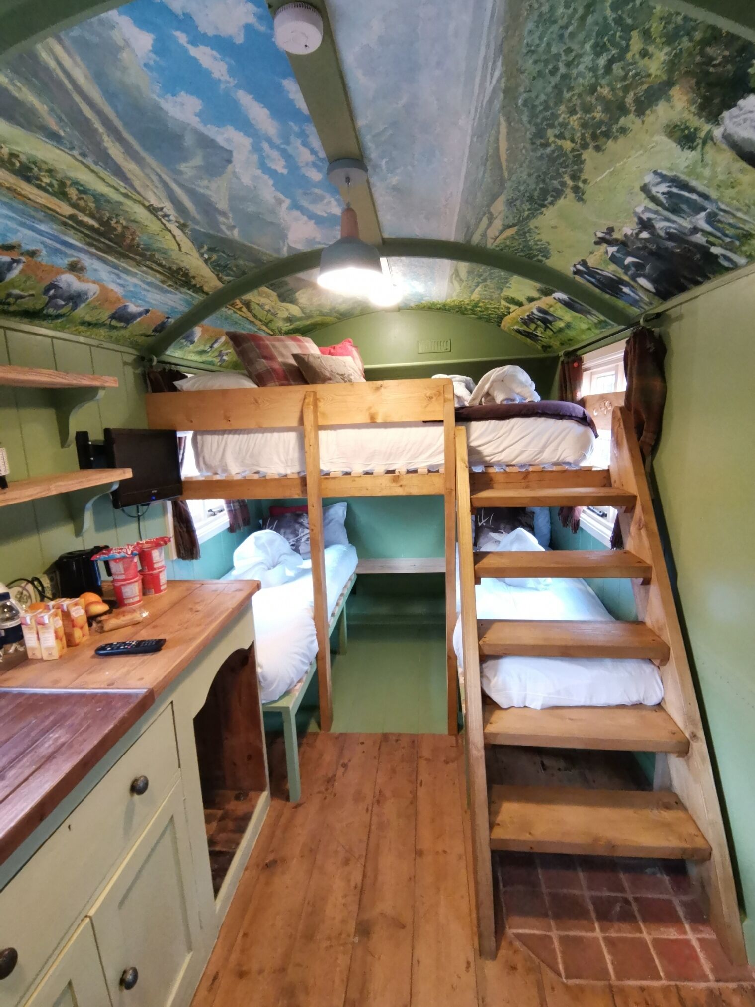 A bunk bed room with a wooden desk and shelves, a ceiling mural, and a staircase.