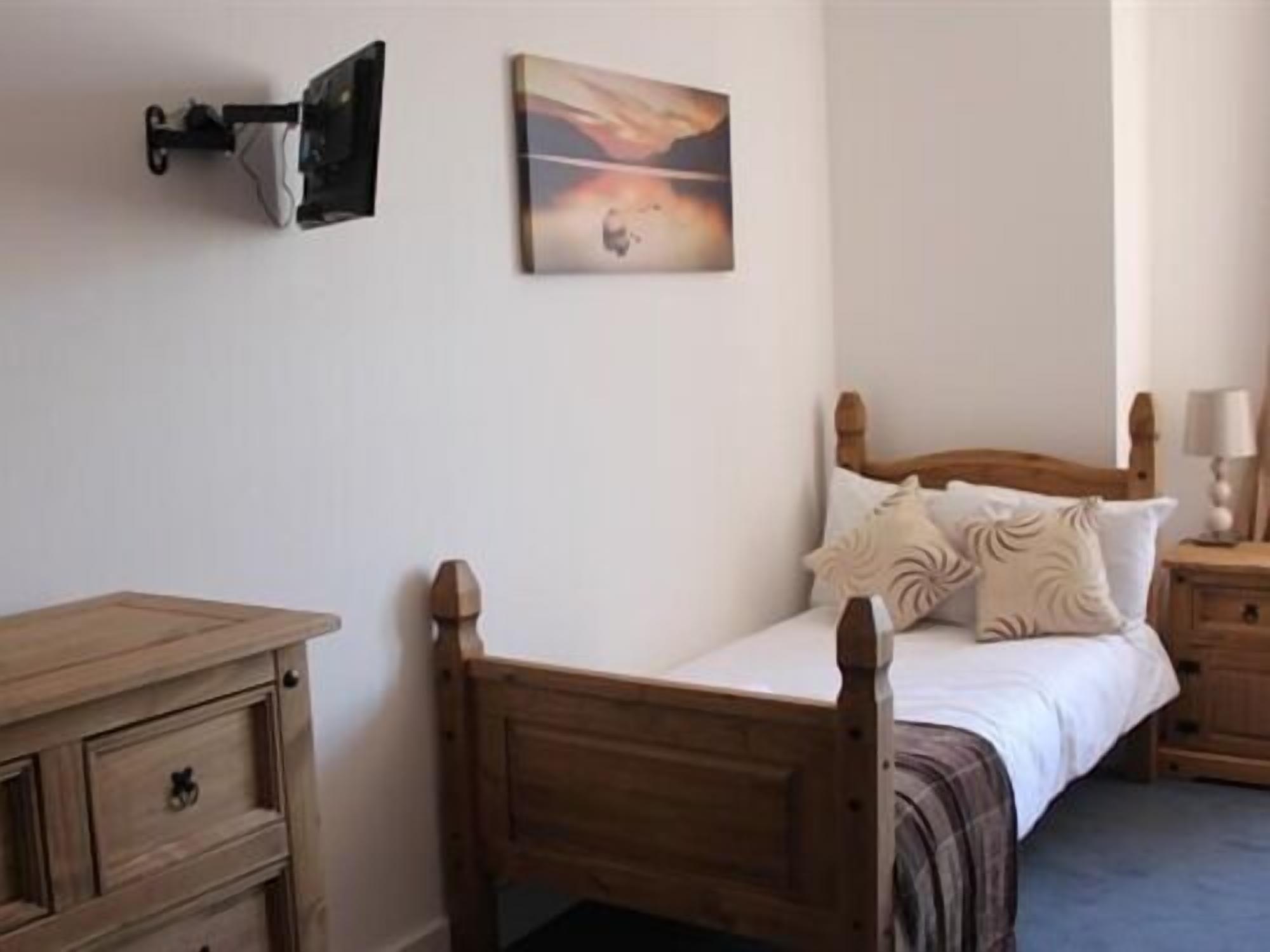 A bedroom with a wooden bed, a nightstand, a dresser, and a wall-mounted lamp.