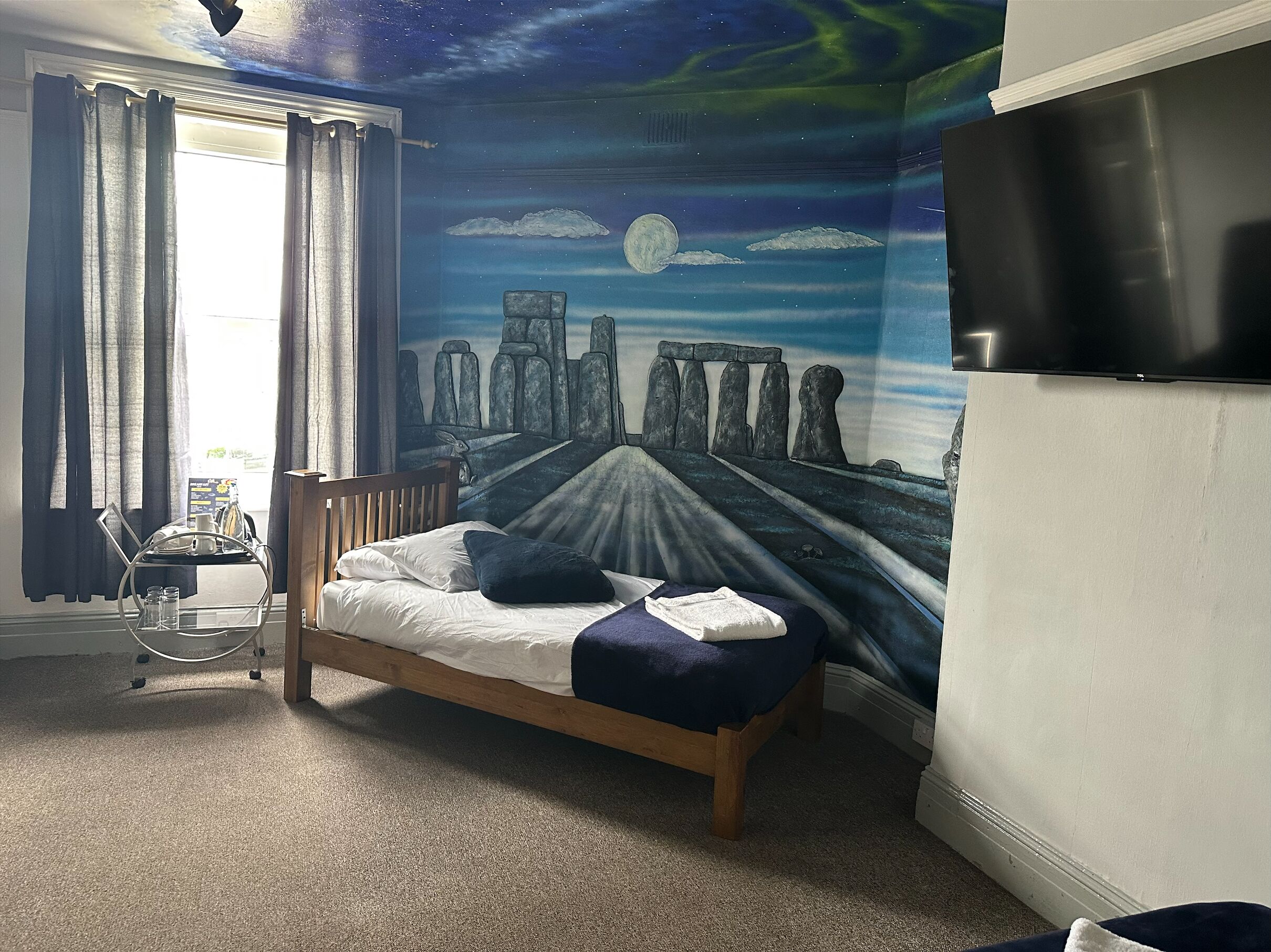 A bedroom with a bed, a night sky mural, a window with curtains, and a small table with a tray.