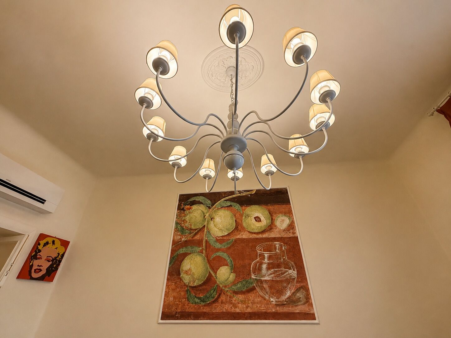 A chandelier with multiple arms and lights, a painting of fruits and a glass pitcher on the wall, and an air conditioning unit on the wall.