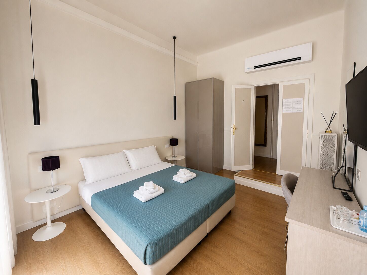 A hotel room with a bed, bedside tables, a desk with a chair, an air conditioning unit, and a television.