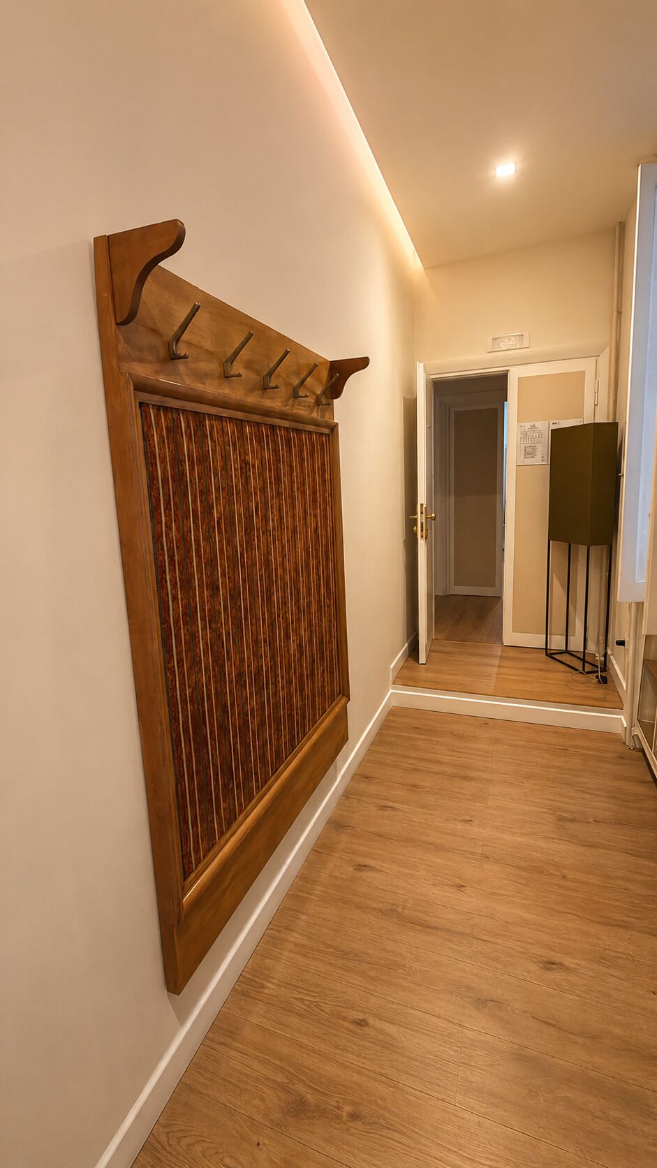 A hallway with wooden flooring, a wooden wall-mounted rack with hooks, and a door leading to another room.