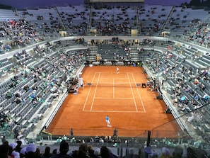 A large indoor tennis stadium with multiple tiers of seating and a clay court.