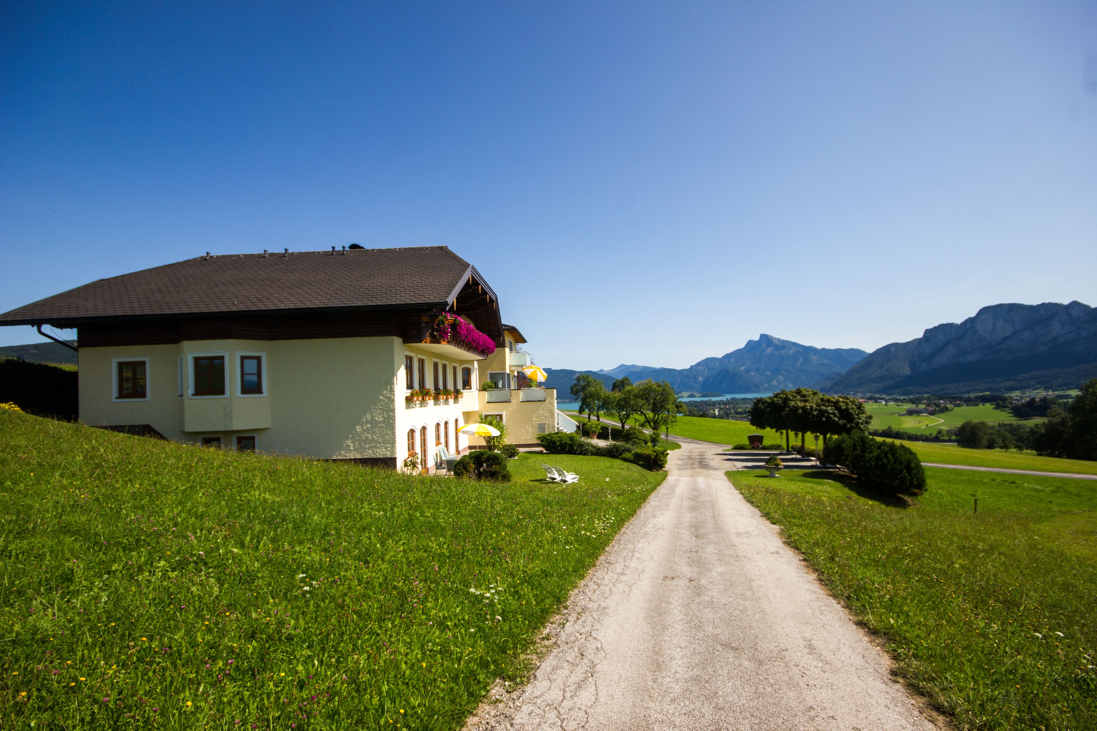 A house with a sloped roof, a balcony with flowers, and a dirt road leading to a mountainous landscape.