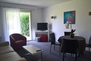A hotel room with a sofa, a small table, a TV, a dining area with chairs, a plant, and a window with curtains. - Hôtel Maison Chiberta (Anglet)