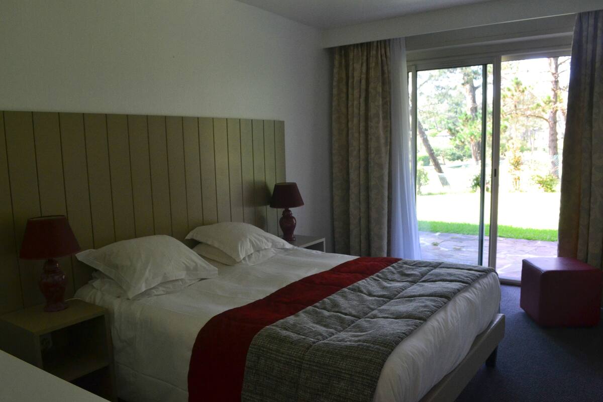 A hotel room with a bed, two bedside lamps, a red accent pillow, a red ottoman, and a view of the outdoors through a sliding glass door.