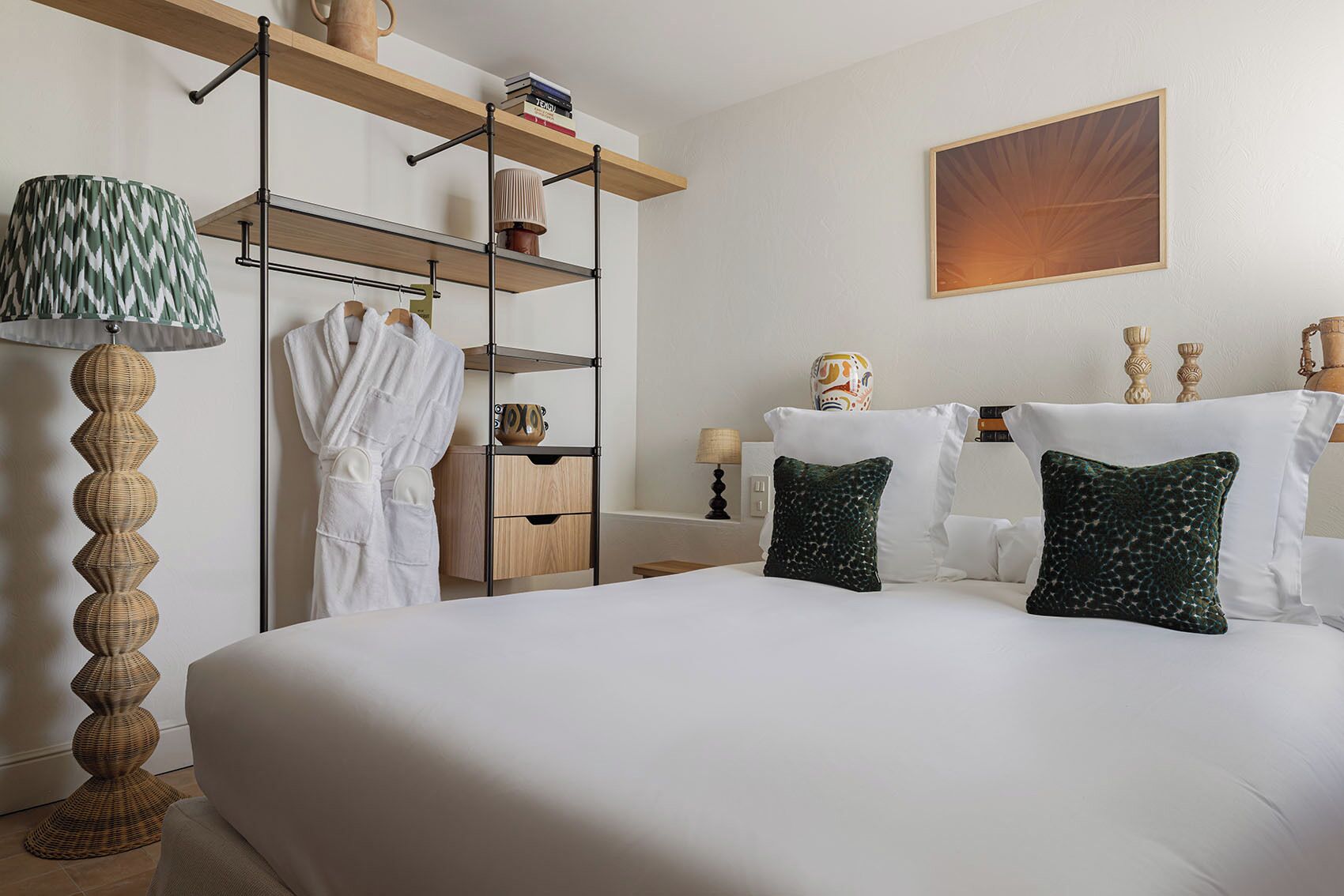 A hotel room with a large bed, a wardrobe with hanging clothes, a lamp, and a painting on the wall.