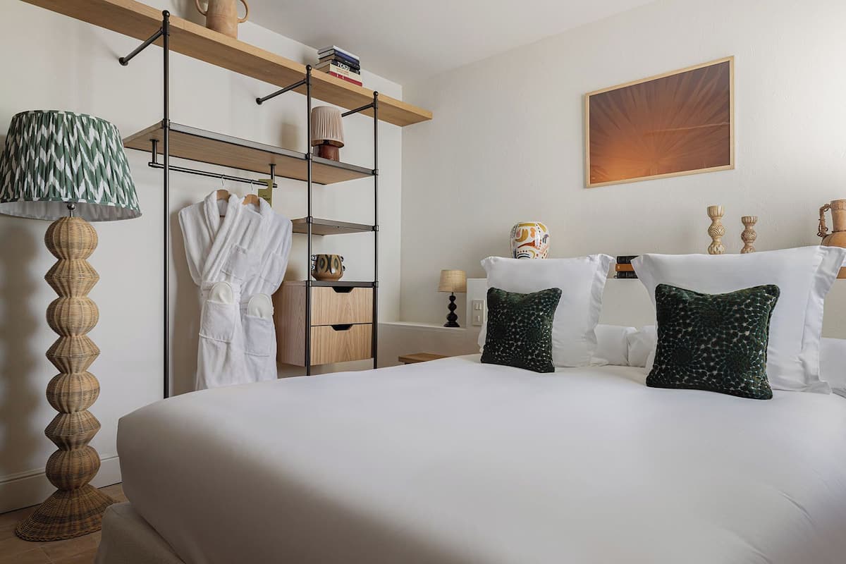 A hotel room with a large bed, a wardrobe with hanging clothes, a lamp, and a painting on the wall.