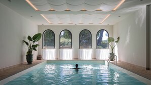 An indoor swimming pool with a person swimming, surrounded by large windows and potted plants. - Hôtel Maison Chiberta (Anglet)