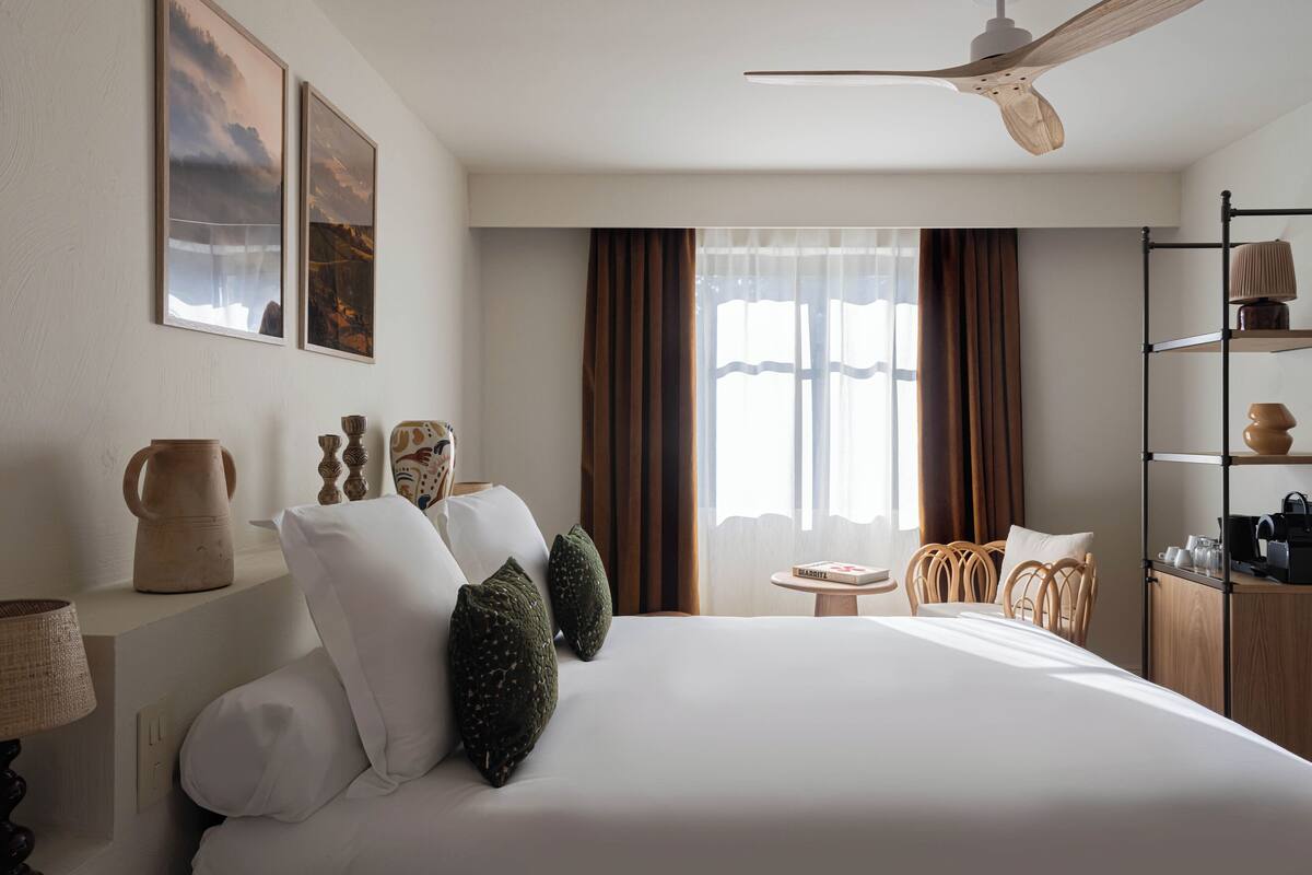 A hotel room with a large bed, a ceiling fan, a small table, and a bookshelf.