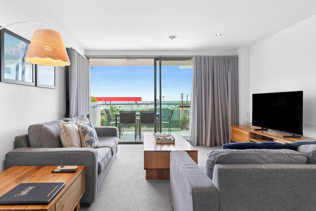 A modern living room with a sofa, a coffee table, a TV, and a balcony with a view.