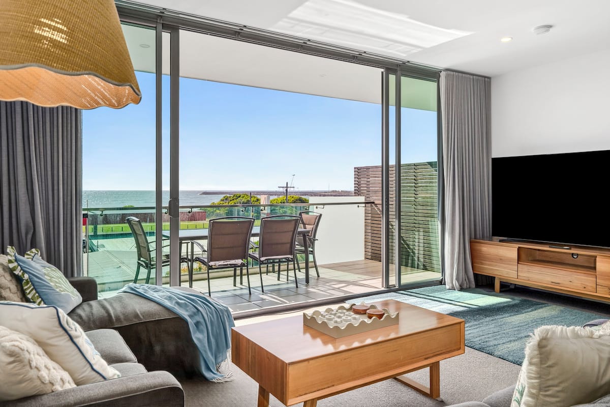 A modern living room with a large TV, a sofa, a coffee table, and a balcony with a view of the sea.