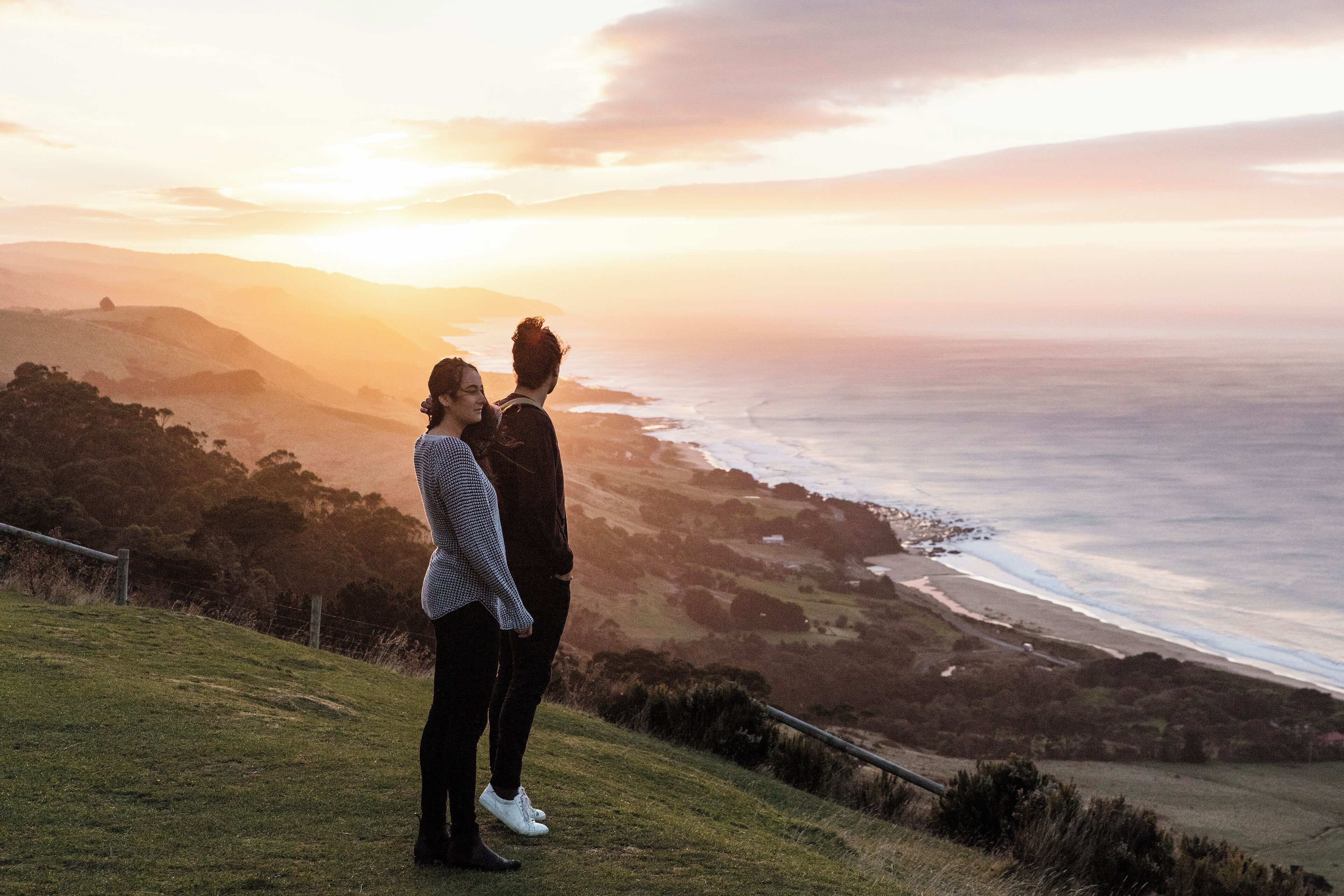 Um casal em pé numa colina gramada com vista para a paisagem costeira ao pôr do sol.