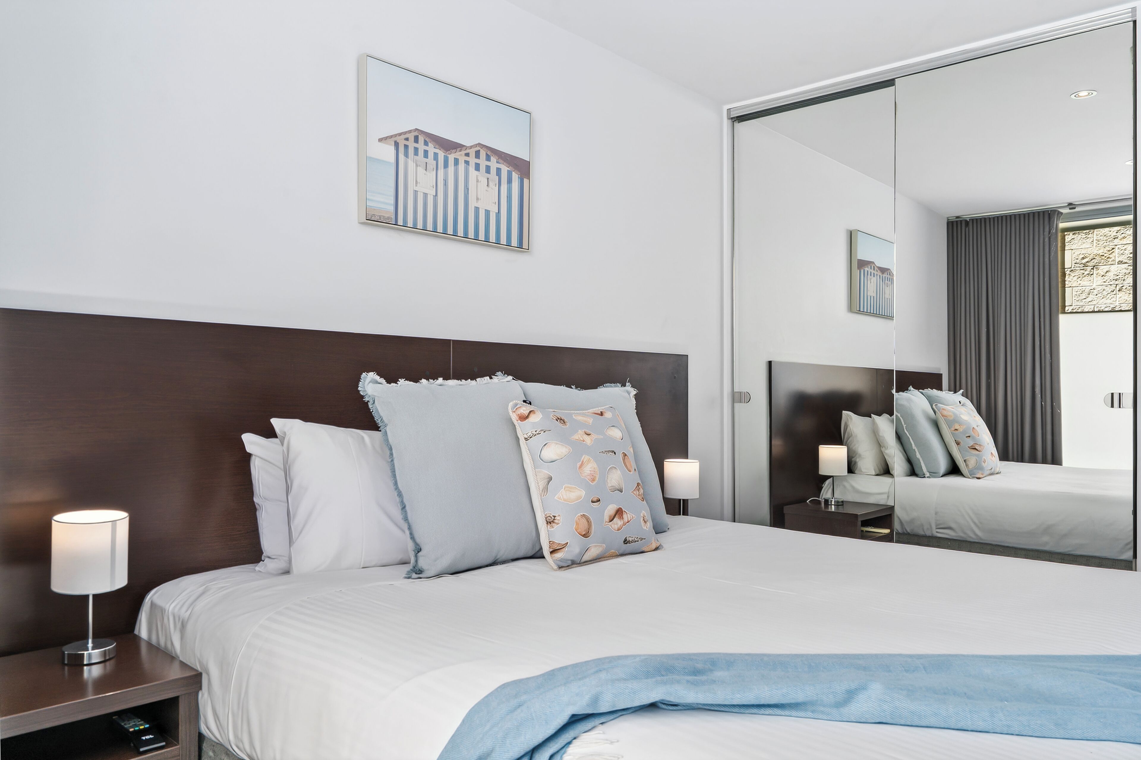 A modern hotel room with a large bed, a nightstand with a lamp, a mirror, and a framed picture on the wall.
