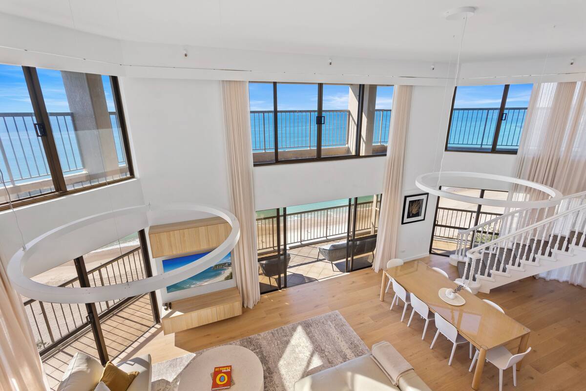 Ampla sala de estar com piso de madeira, uma mesa de centro redonda e uma mesa de jantar com cadeiras. Há uma grande janela com vista para o mar e uma varanda com mobiliário para área externa.