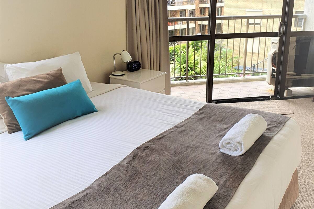 Quarto de hotel com uma cama grande, uma mesa de cabeceira com luminária, vista para a varanda e uma janela com cortinas.