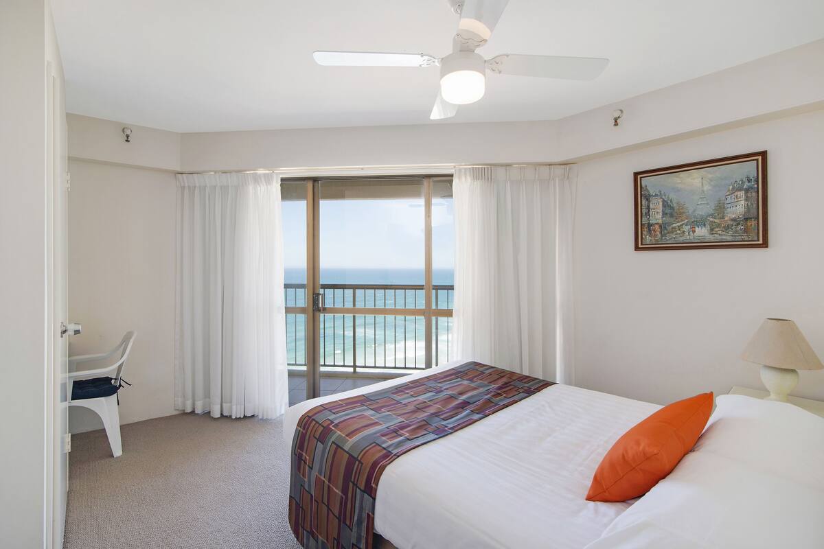 Quarto de hotel com cama, escrivaninha, cadeira, vista para o mar e um quadro na parede.
