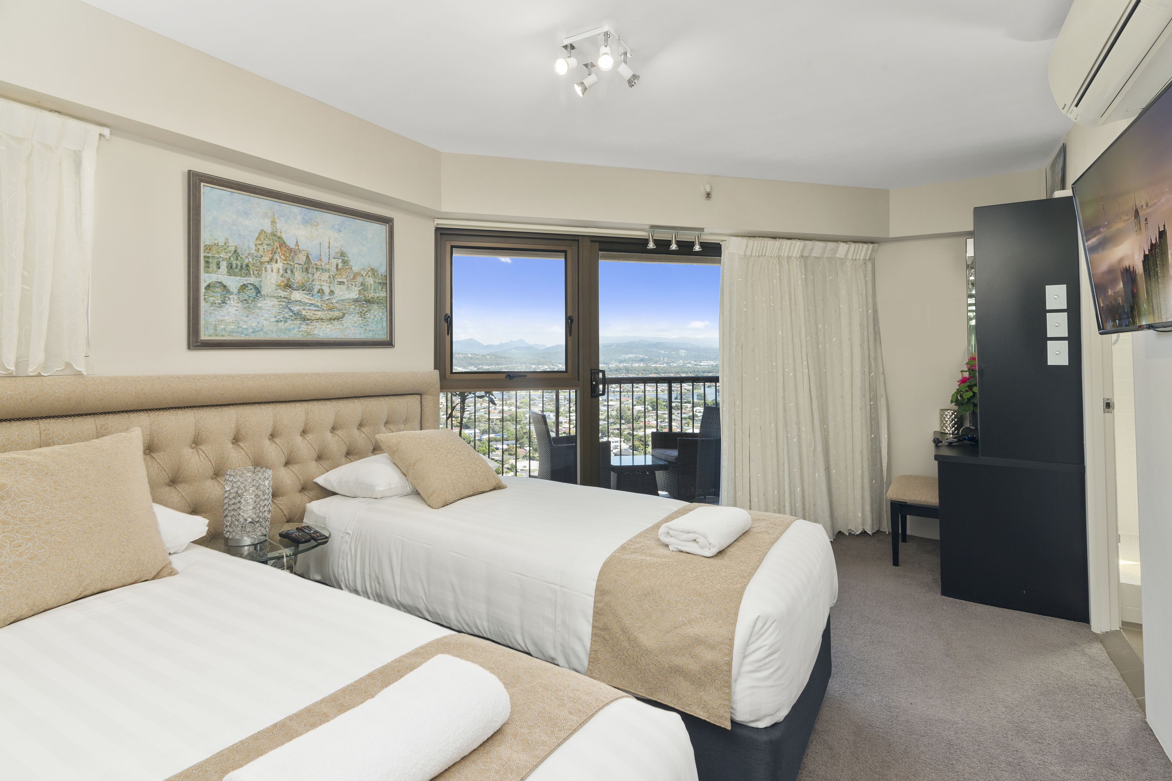 Camera d'albergo con due letti, un'ampia finestra vista città, un balcone con tavolo e sedie e una televisione a parete.