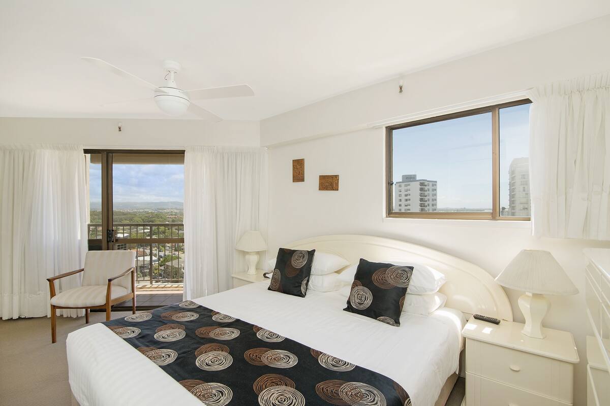 Quarto de hotel com uma cama grande, uma poltrona, uma mesa de cabeceira, um abajur e vista da cidade através de uma grande janela.