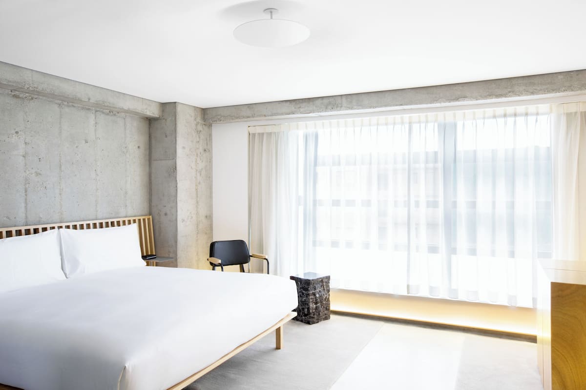 A modern hotel room with a large bed, a chair, a small table, and a window with sheer curtains.