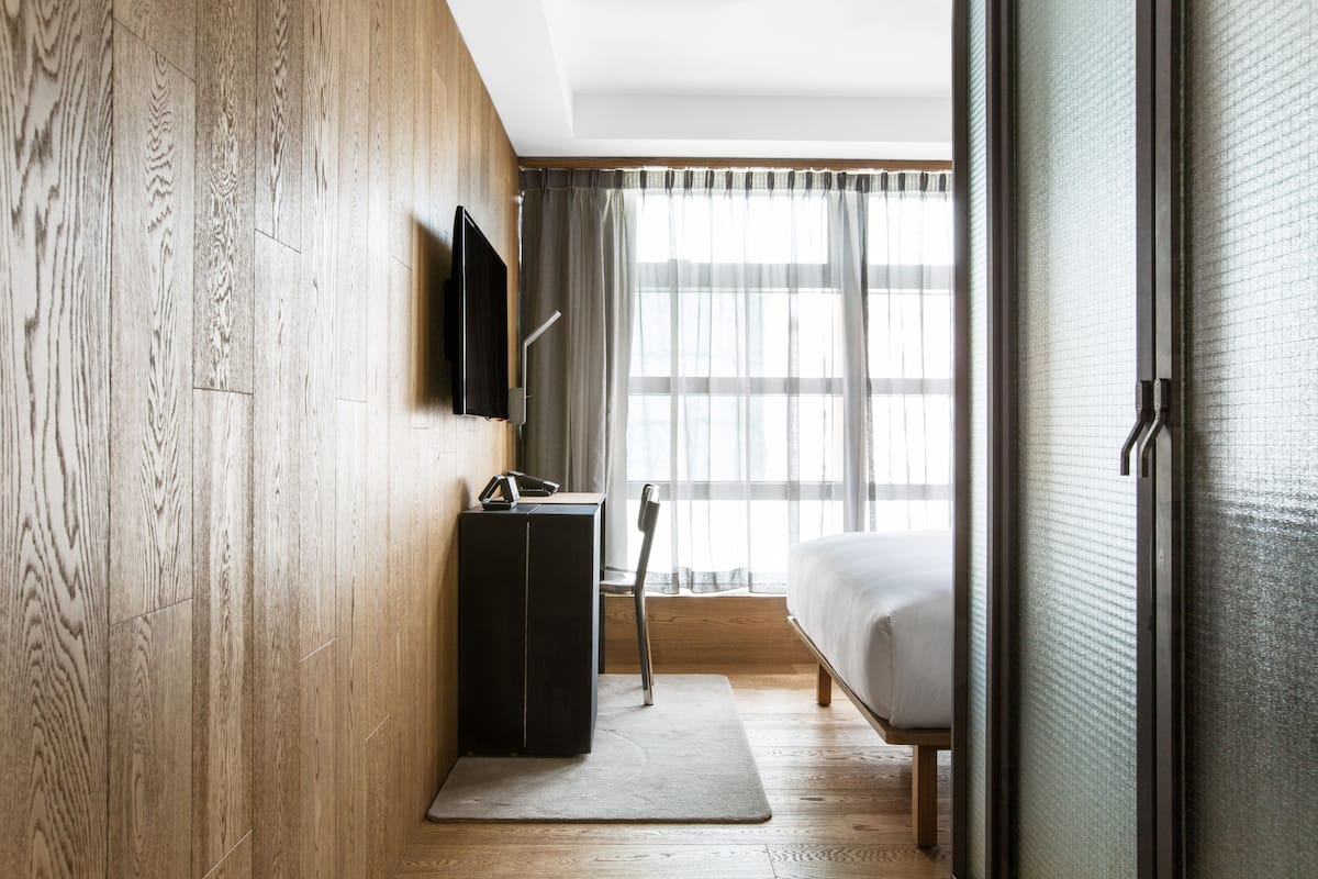 A hotel room with a wooden wall, a flat-screen TV, a desk with a chair, a bed, and a large window with sheer curtains.