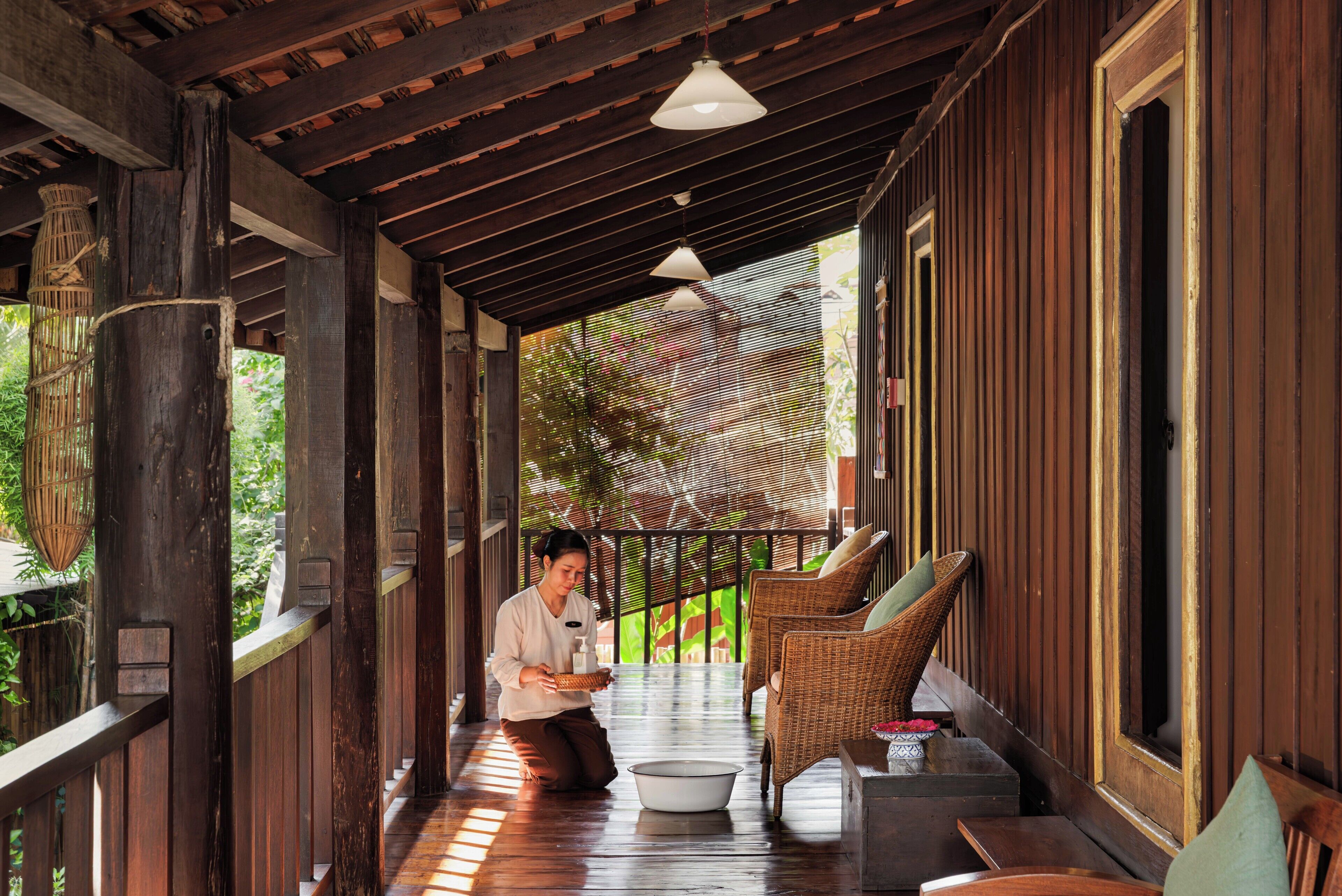 A wooden porch with a person kneeling, a wicker chair, and a white basin.