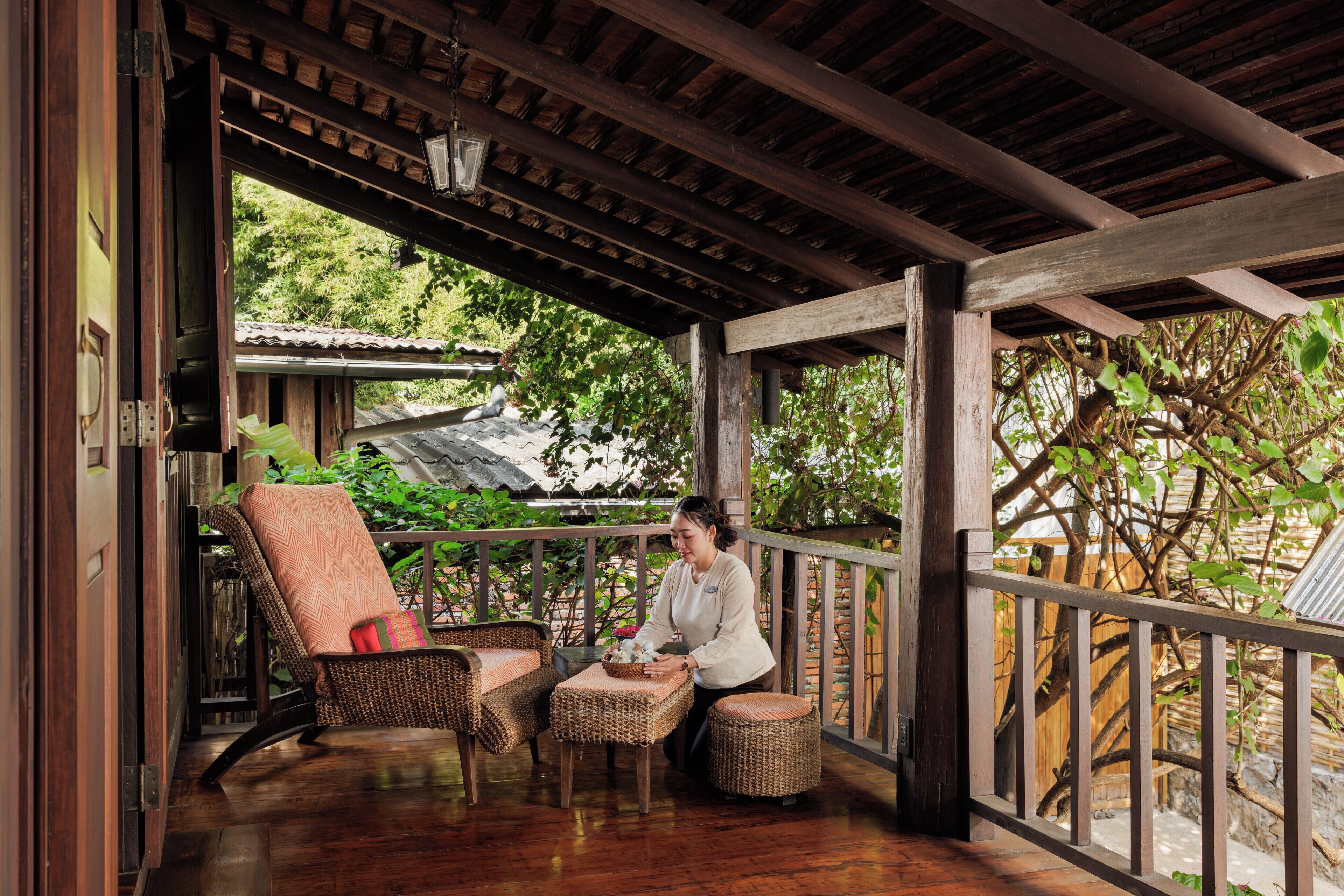 A wooden porch with wicker furniture, a table, and a view of a traditional house and trees.