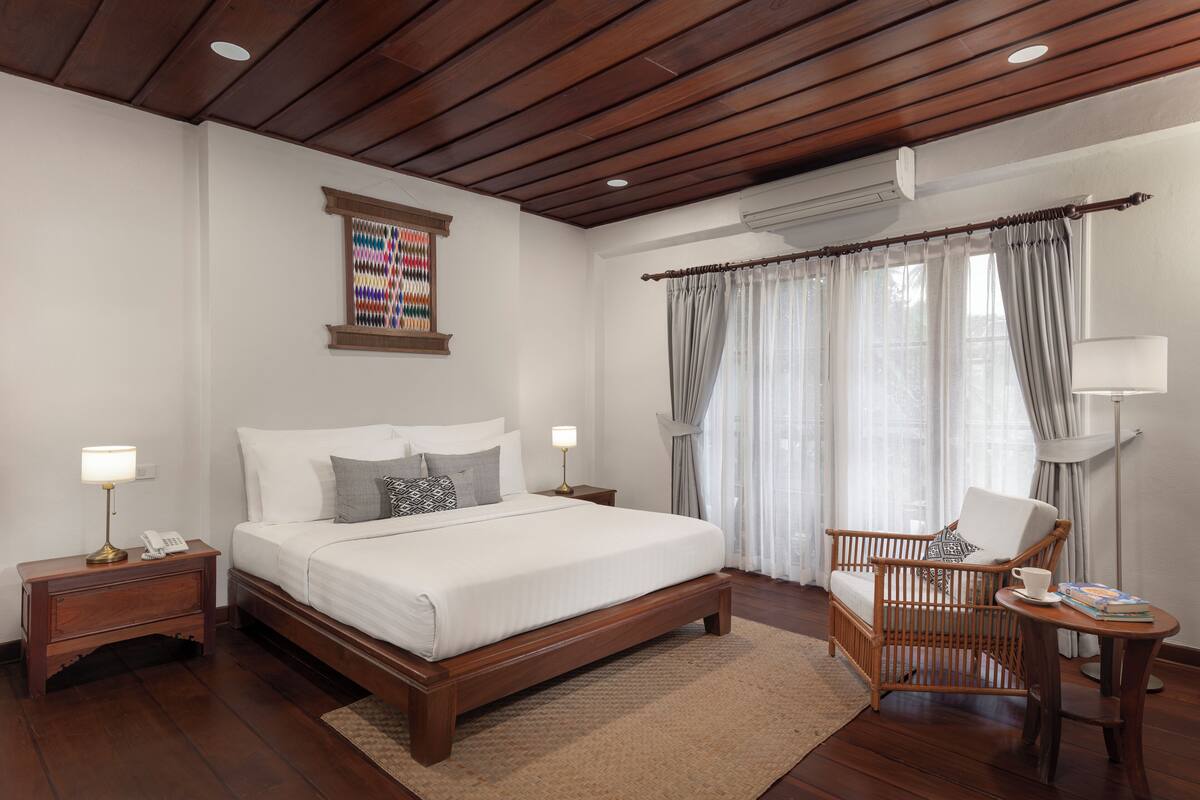 Un dormitorio con cama de madera, mesitas de noche, una silla de mimbre, una lámpara de pie y un ventanal con cortinas.