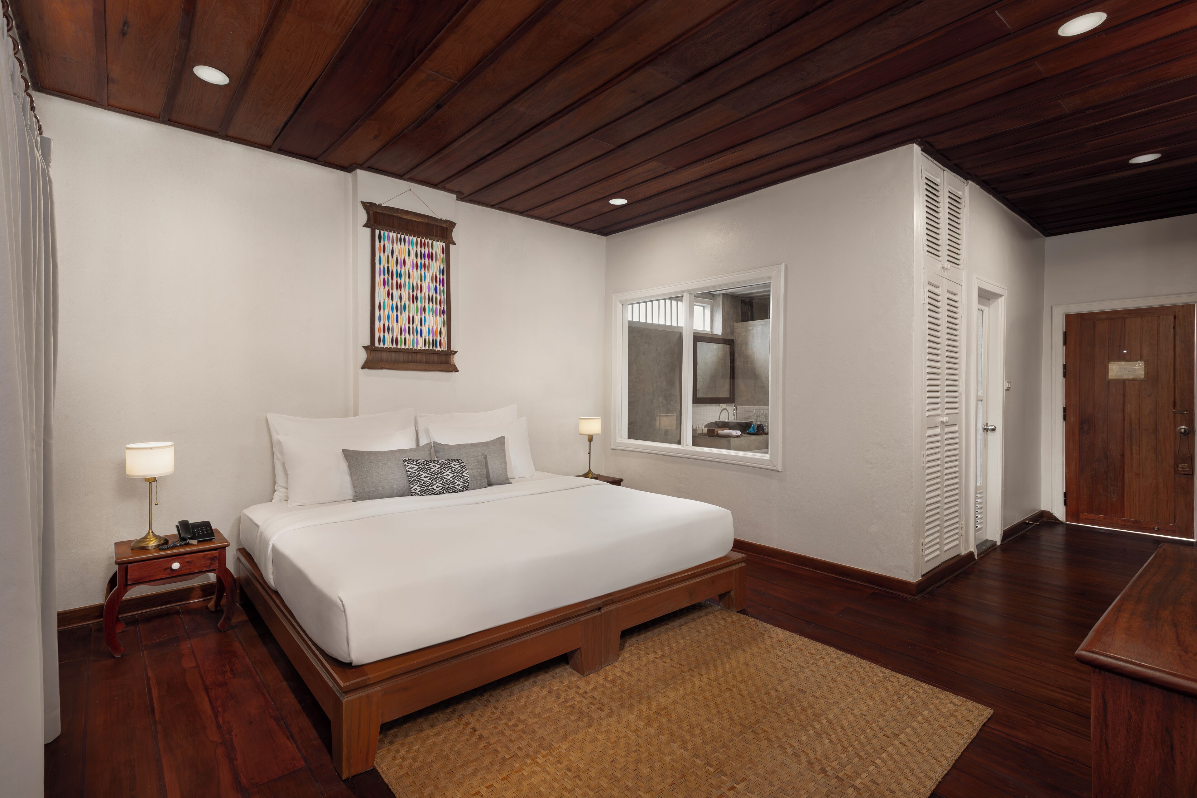 A bedroom with a wooden bed frame, white bedding, a small bedside table with a lamp, a window with blinds, and a wooden door.