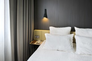 Habitación de hotel con cama, almohadas blancas, mesita de noche con vaso de agua, lámpara y velador con un libro y control remoto.