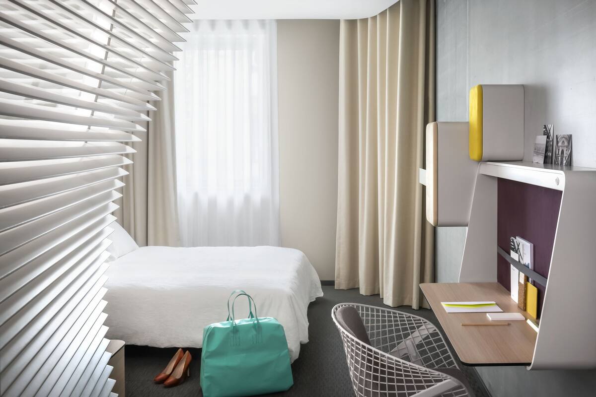 A modern hotel room with a bed, a desk with a chair, a window with blinds, and a small bookshelf.