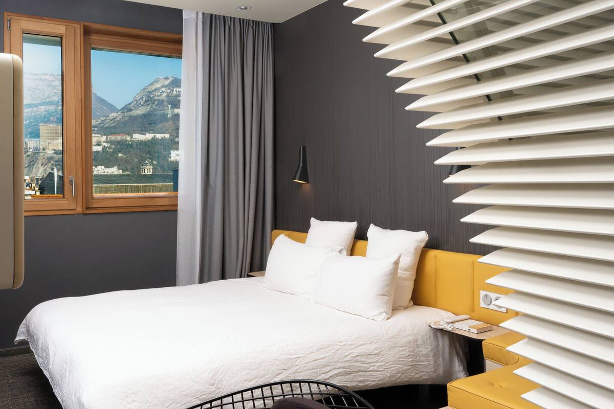 A hotel room with a bed, a nightstand, a lamp, and a window with a view of mountains.