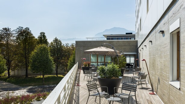 A balcony with outdoor seating, a large potted plant, and a view of a modern building and trees. - OKKO Hotels Grenoble Centre (Grenoble)