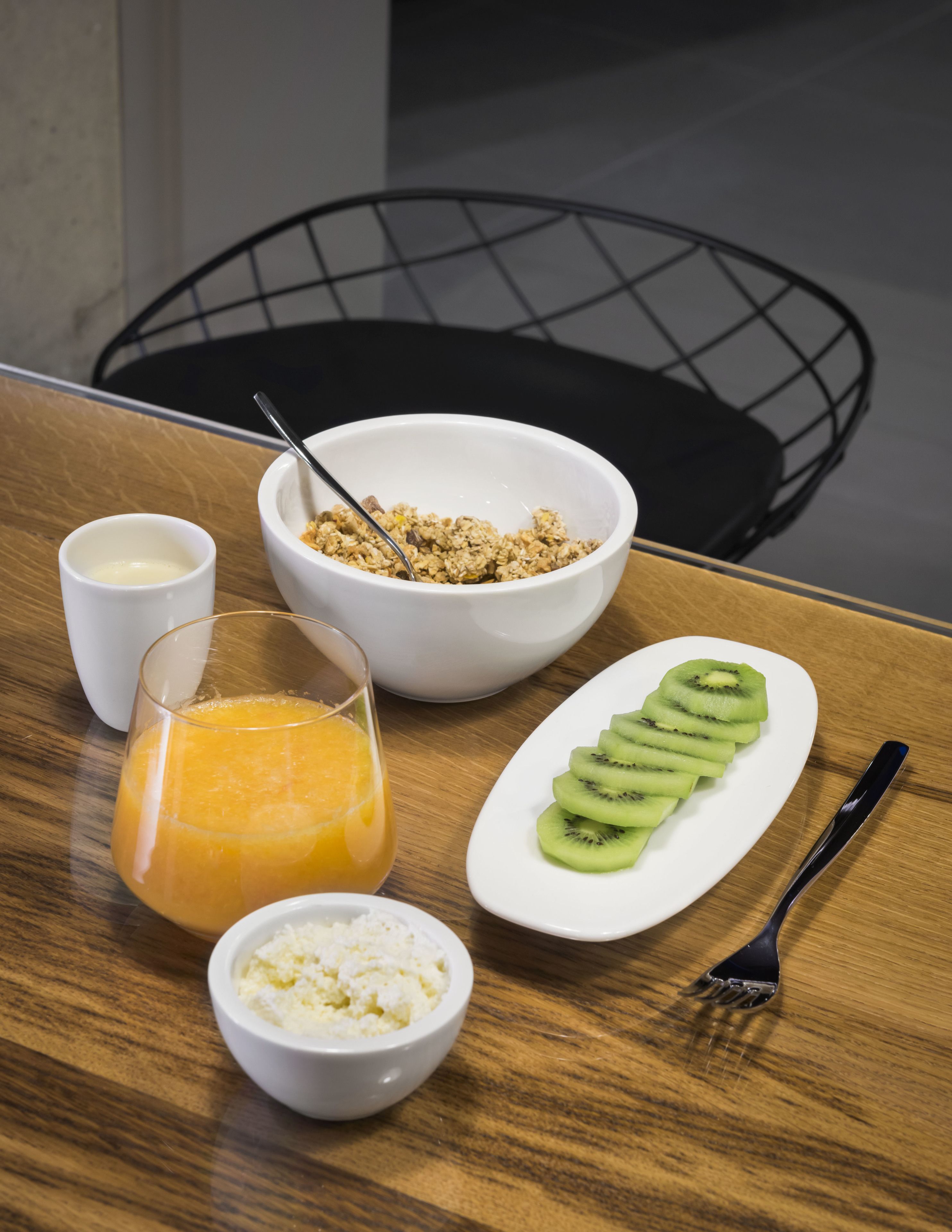 Ein Frühstückstisch mit einer Schüssel Müsli, einem Glas Orangensaft, einem Teller mit Kiwischnitten, einer kleinen Schüssel mit Hüttenkäse und einer Milchbecher.