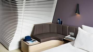 Una habitación de hotel moderna con un sofá curvo, una mesita de noche con un libro y un sombrero, una lámpara y un ventanal grande con persianas.