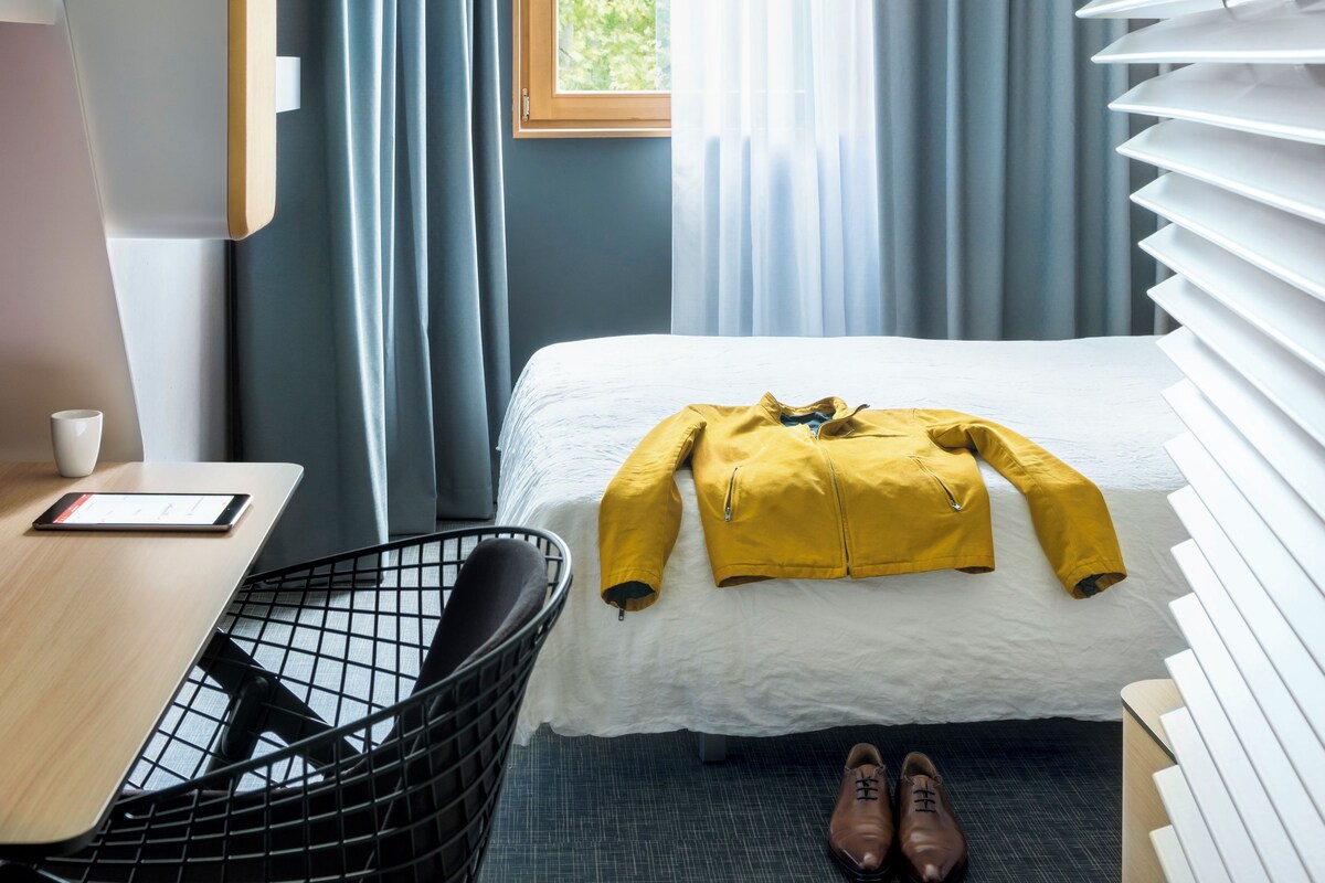 Habitación de hotel con cama, un escritorio con silla, una ventana con cortinas y una lámpara de pared.
