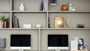 A modern office space with two desks, each with a computer monitor, keyboard, and mouse. There are two black office chairs with wheels. The room has a grey wall and shelves with various decorative items and books. - OKKO Hotels Grenoble Centre (Grenoble)