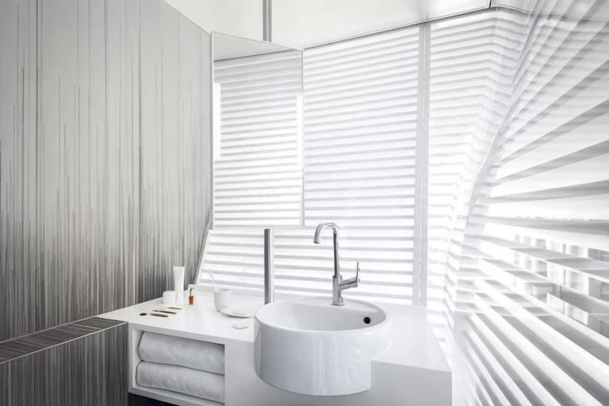 Une salle de bain moderne avec un lavabo blanc, un miroir et une fenĂȘtre avec des stores.